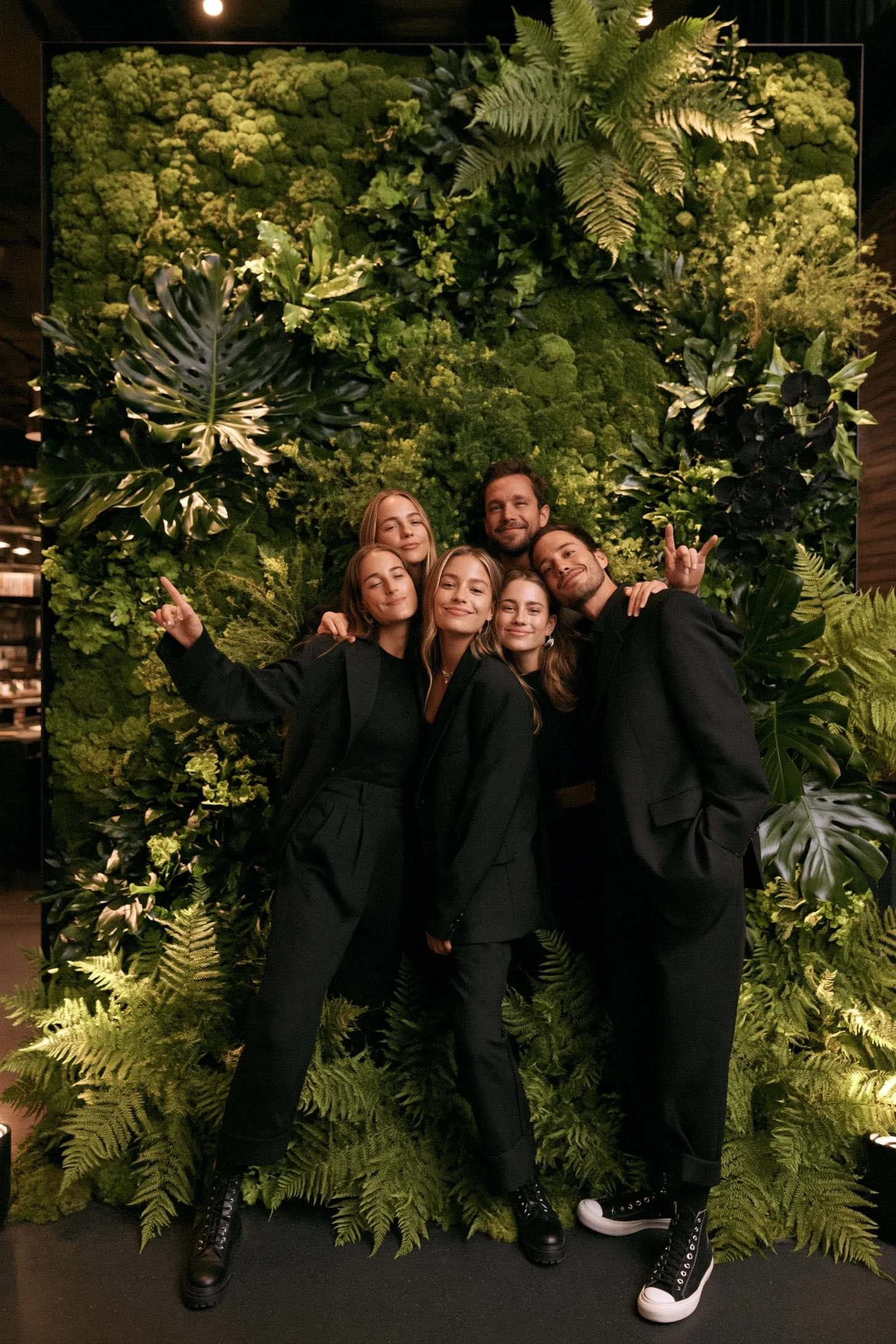 Wedding guests posing in front of a lush fern and white daisy photo booth backdrop at night