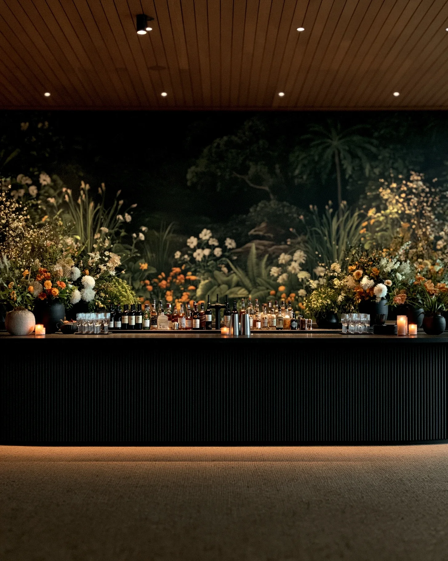 A statement island bar at a nighttime wedding, draped in ferns and foliage with warm low lighting